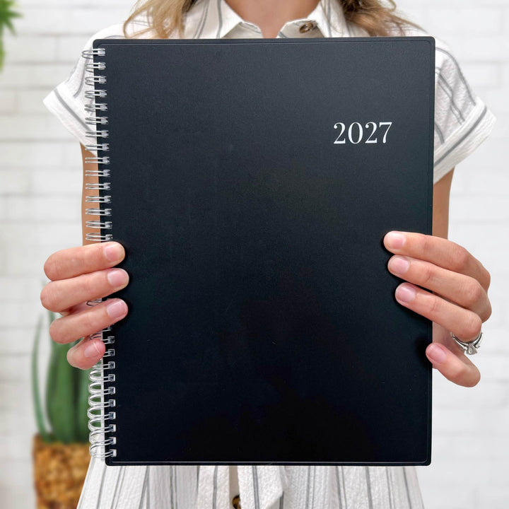 A person holding a black 2027 spiral-bound planner in front of their face, with a white brick wall and a potted plant in the background.