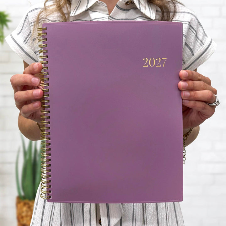 A person holding a purple 2027 spiral-bound planner in front of their face, wearing a white and gray striped shirt against a white brick wall background.
