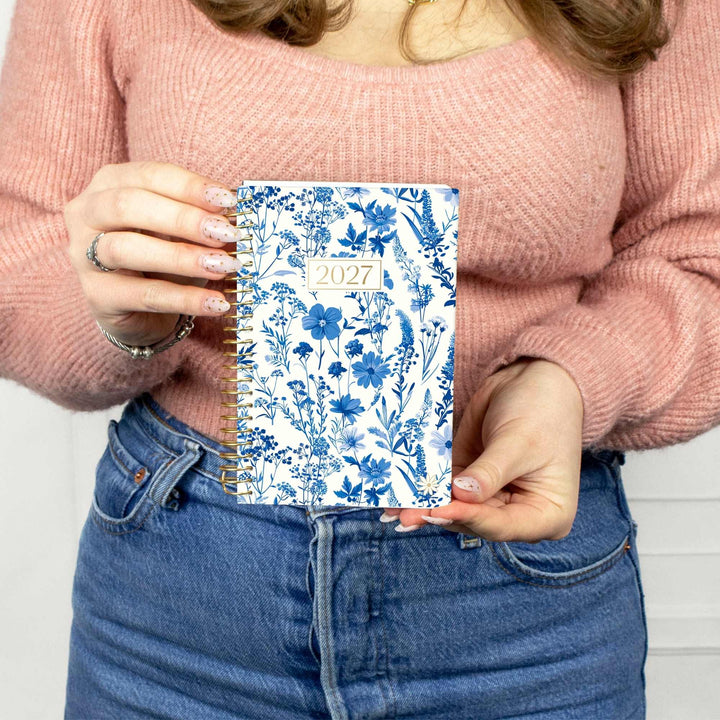 A person in a pink knit sweater and blue jeans holds a 2027 spiral-bound planner with a blue and white floral pattern.