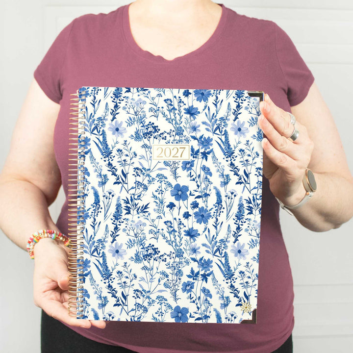 A person in a mauve t-shirt holds up a 2027 spiral-bound planner with a blue and white floral patterned cover against a white paneled wall.