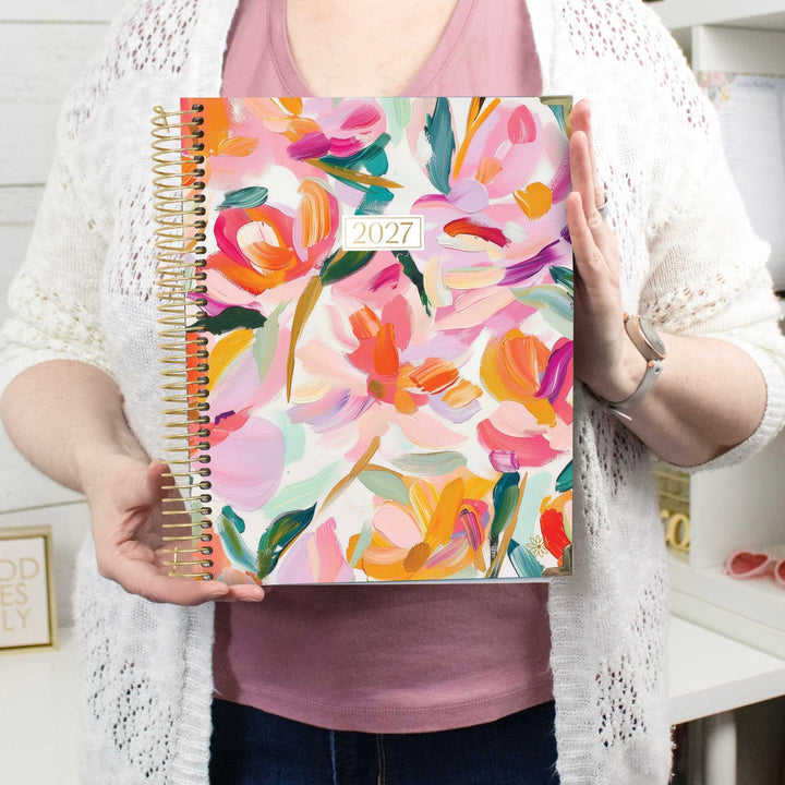 A person holds a 2027 spiral-bound planner with a colorful floral cover. The planner features a vibrant painting of flowers in pink, orange, yellow, and green on a white background.