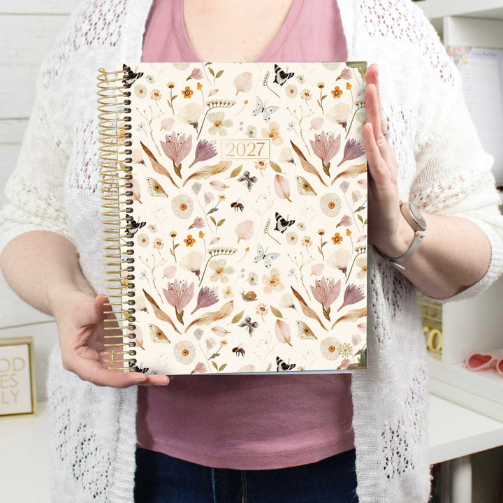 A person holding a 2027 spiral-bound planner with a cream floral cover. The planner features a pattern of pink and purple flowers, white blossoms, and small butterflies and bees on a cream background, with a gold-toned spiral binding.