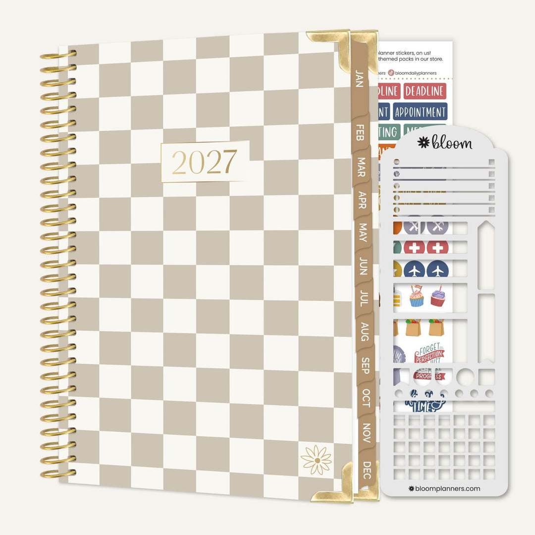 A 2027 spiral-bound planner with a beige and white checkerboard cover, shown with a gold page divider and a white sticker sheet on a light gray background. The sticker sheet includes the 'bloom' brand logo and various colorful planner stickers.