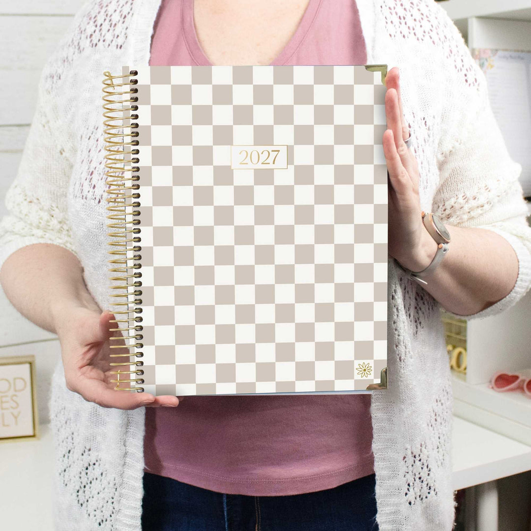 A person holding a 2027 spiral-bound planner with a beige and white checkerboard cover. The planner features a gold wire coil binding and a small gold sunflower logo in the bottom right corner.