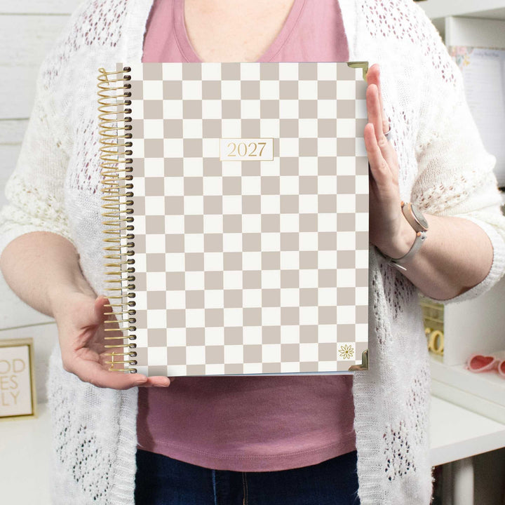 A person holding a 2027 spiral-bound planner with a beige and white checkerboard cover. The planner features a gold wire coil binding and a small gold sunflower logo in the bottom right corner.