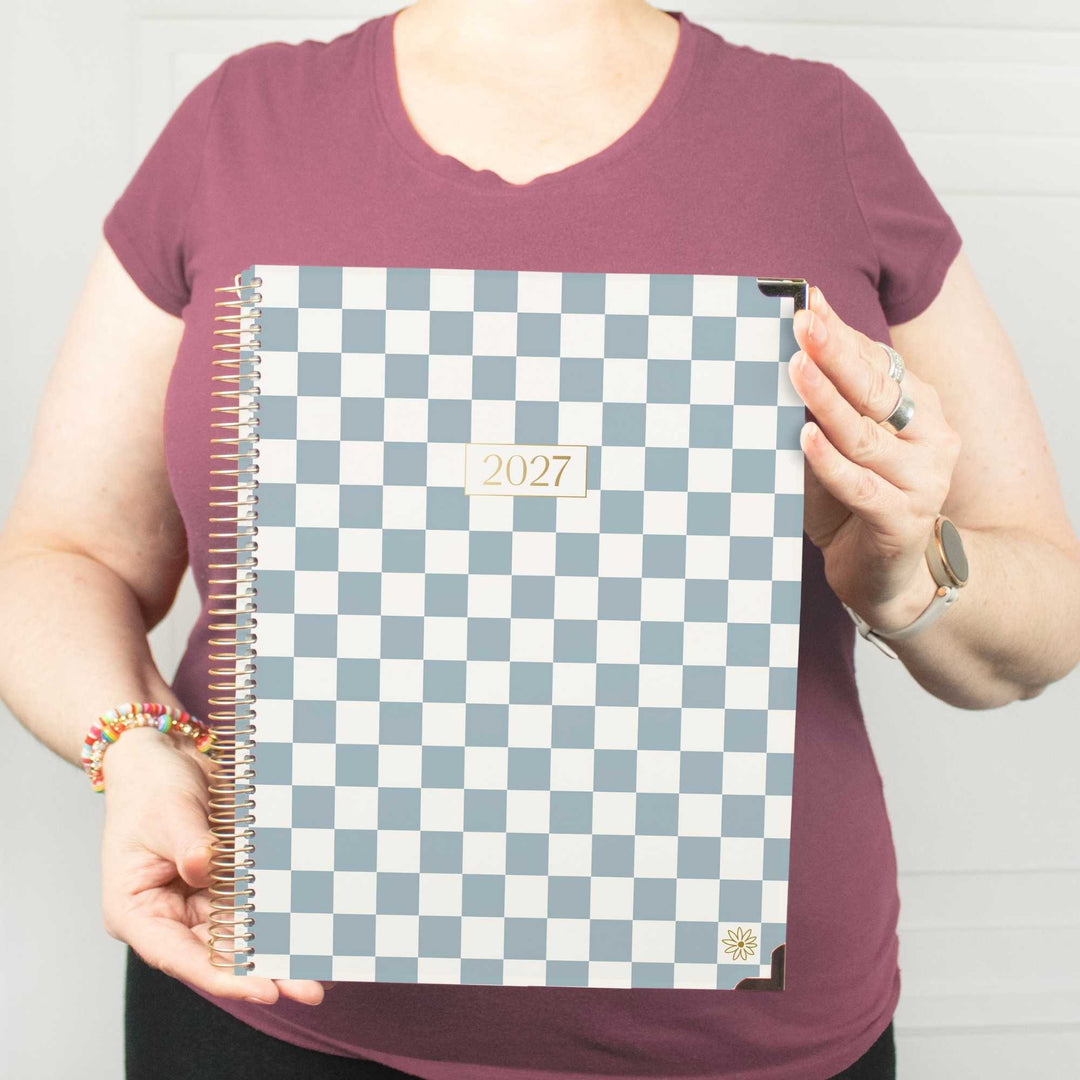 A person in a mauve t-shirt holds up a 2027 spiral-bound planner with a blue and white checkerboard cover.