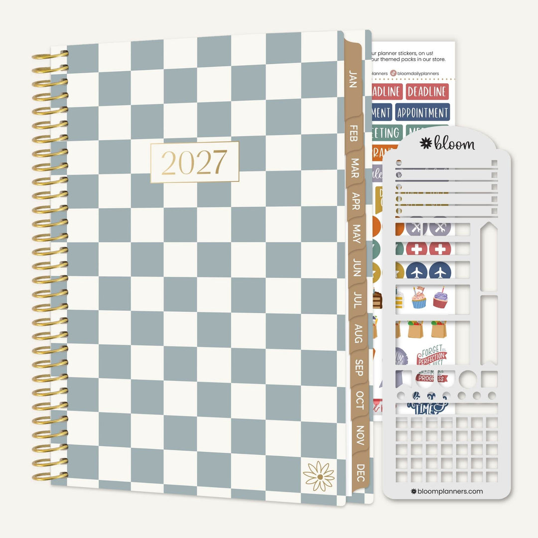 A 2027 spiral-bound planner with a blue and white checkerboard cover, shown alongside a white sticker sheet with colorful icons and labels. The planner has a vertical tab with months in gold text and a small gold floral emblem near the bottom.