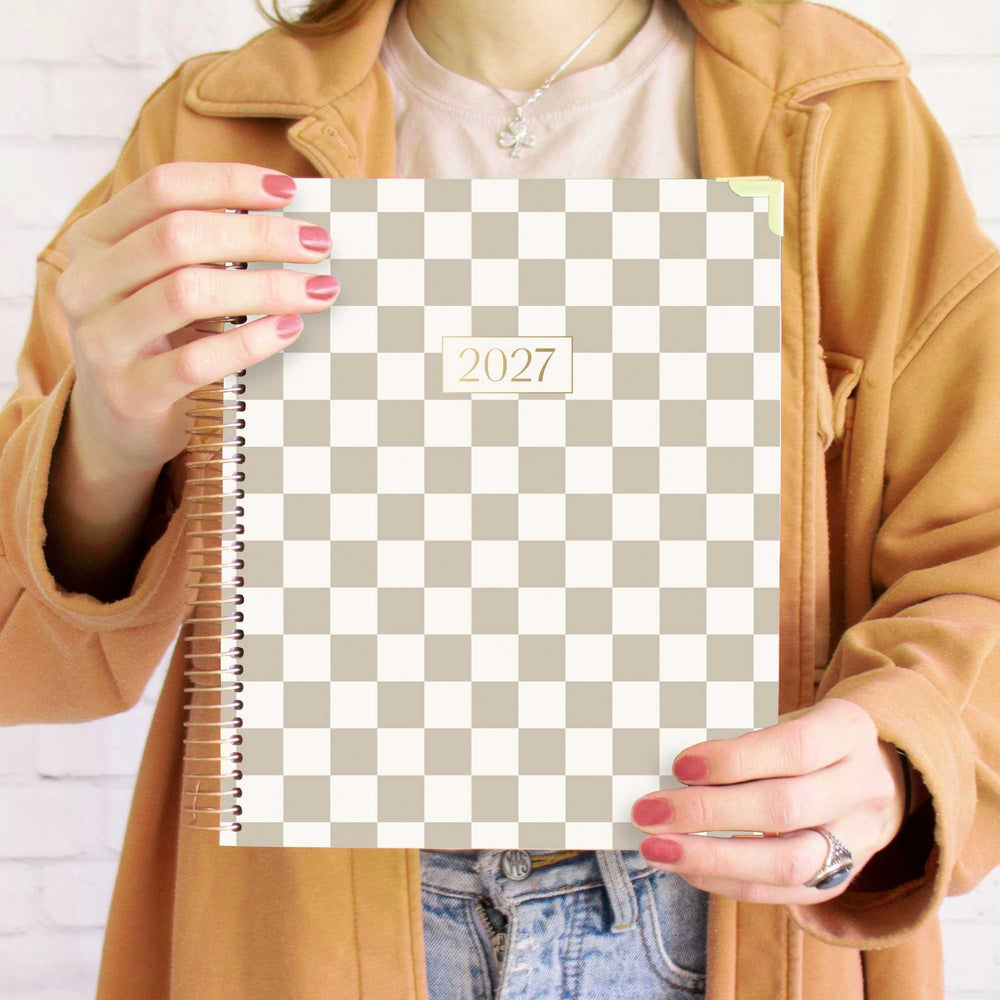 A person holding a 2027 spiral-bound planner with a beige and white checkerboard pattern. The person is wearing a tan jacket, a light pink t-shirt, and blue jeans, with red painted fingernails. - hover