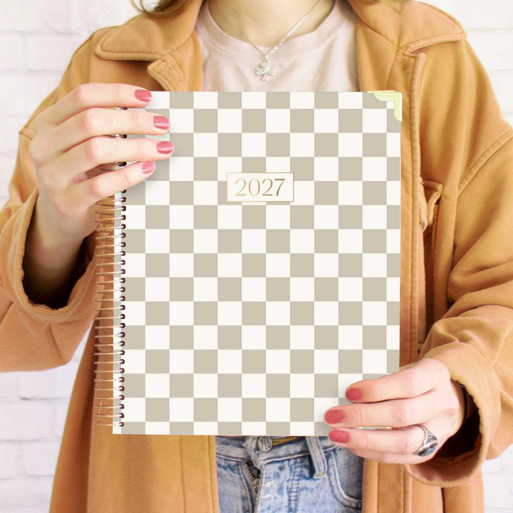 A person holding a 2027 spiral-bound planner with a beige and white checkerboard pattern. The person is wearing a tan jacket, a light pink t-shirt, and blue jeans, with red painted fingernails.