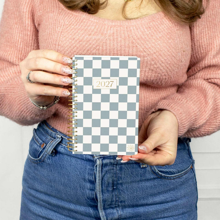 A person in a pink knit sweater and blue jeans holds a spiral-bound 2027 planner with a blue and white checkerboard pattern.