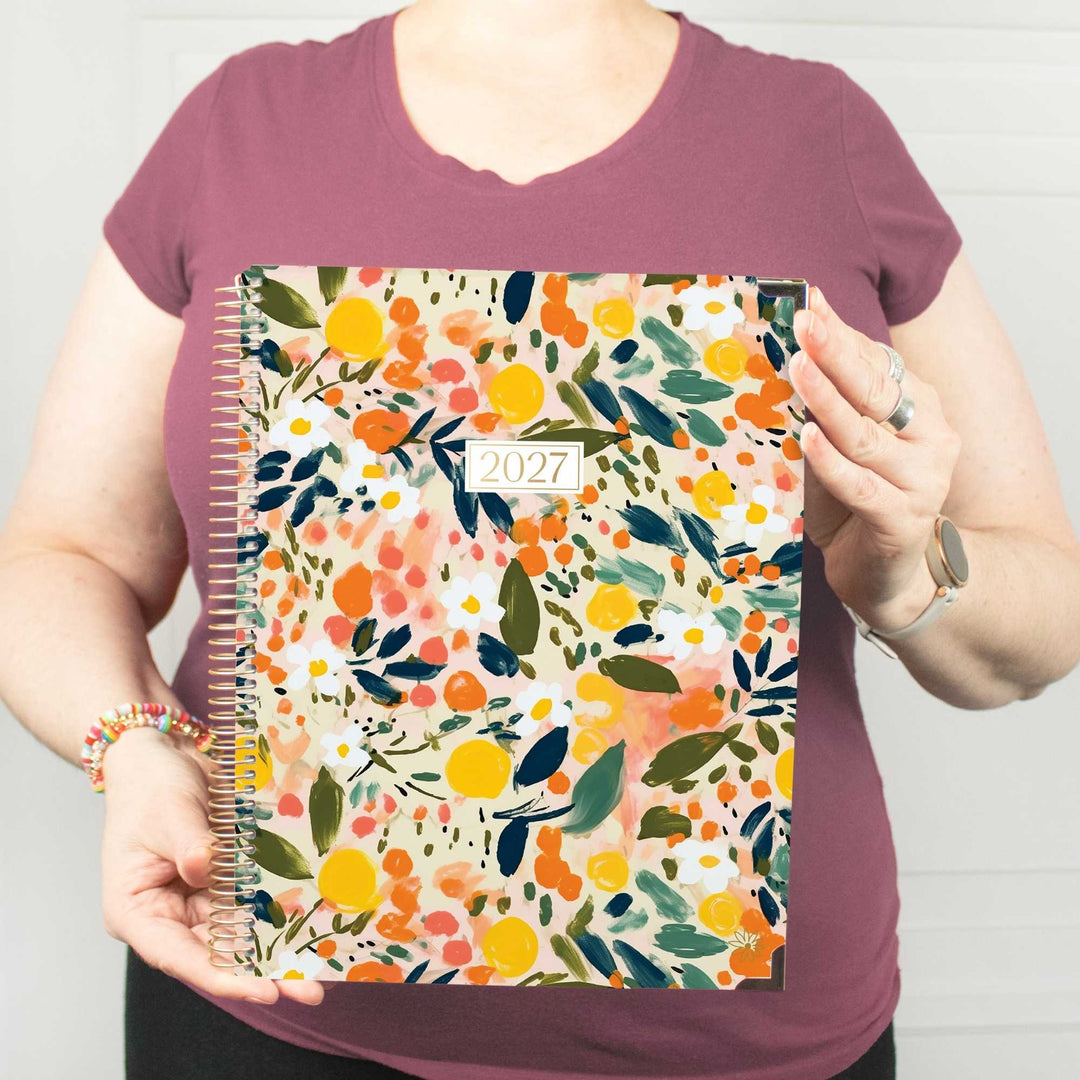 A person wearing a mauve t-shirt holds up a 2027 spiral-bound planner with a colorful watercolor floral pattern featuring oranges, yellows, and greens on a cream background.