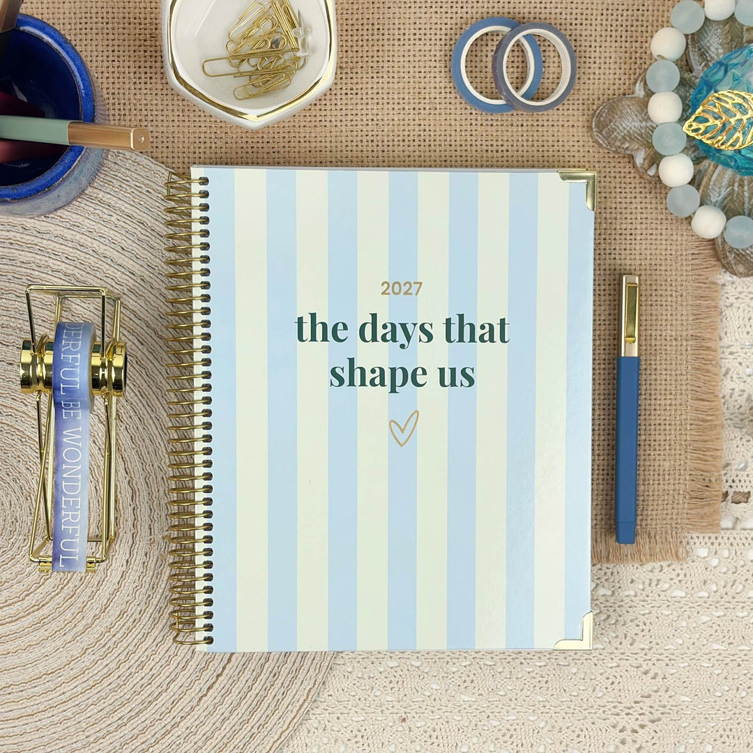 A spiral-bound planner or journal cover laid flat on a woven mat, with the text '2027 the days that shape us' in dark teal and gold. The cover has pale blue and white vertical stripes.
