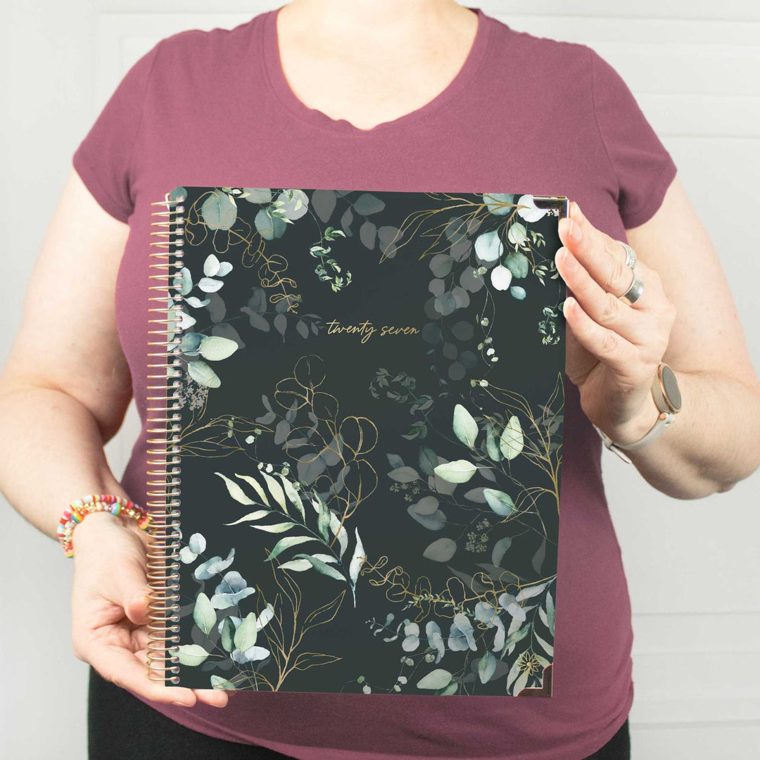 A person wearing a mauve t-shirt holds up a spiral-bound planner with a dark green and black botanical pattern featuring gold leaf details and the text 'twenty seven'.