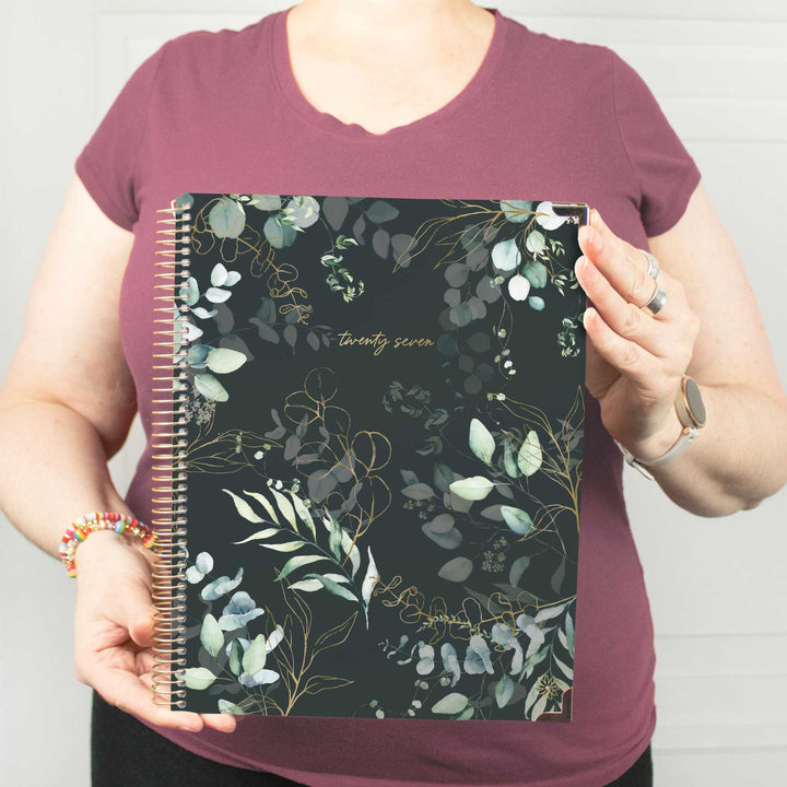 A person wearing a mauve t-shirt holds up a spiral-bound planner with a dark green and black botanical pattern featuring gold leaf details and the text 'twenty seven'.