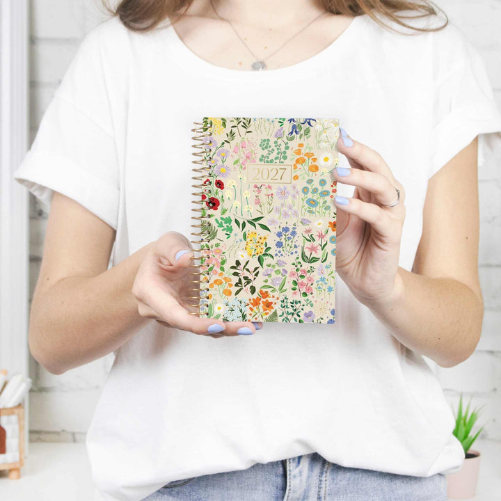 A woman in a white t-shirt holds up a spiral-bound 2027 floral planner against a bright, light-colored background.