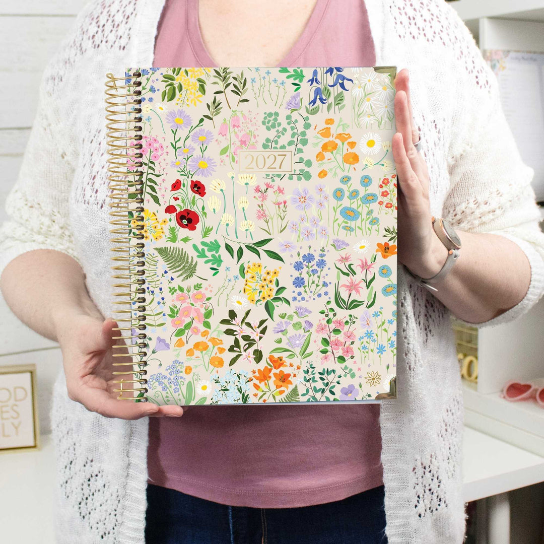 A person holding a 2027 spiral-bound planner with a colorful floral pattern on the cover. The person is wearing a pink top, a white open-knit cardigan, and blue jeans, standing in a bright indoor setting.