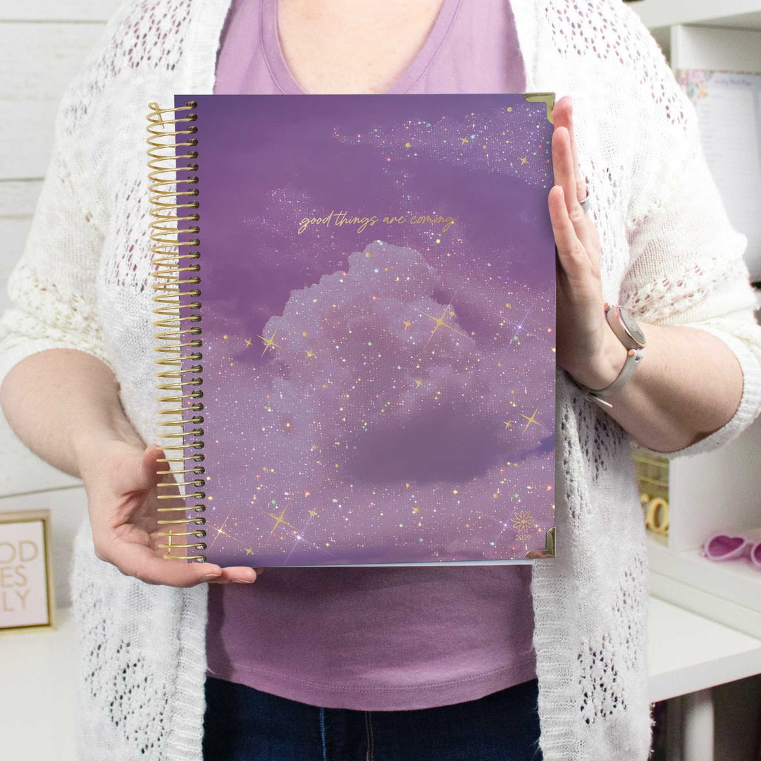 A person holding a purple spiral-bound notebook with a galaxy cloud cover and gold foil text. The person is wearing a white knit cardigan over a purple shirt.