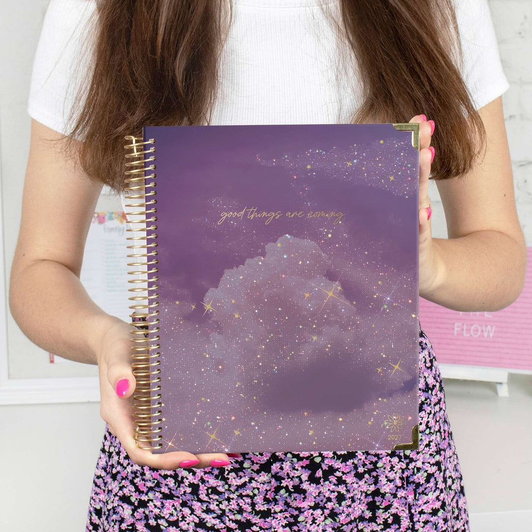 A person holding a purple spiral-bound planner or notebook with a gold glitter cloud design and the phrase 'good things are coming' in gold script. The person is wearing a white t-shirt and a black floral skirt with pink flowers.