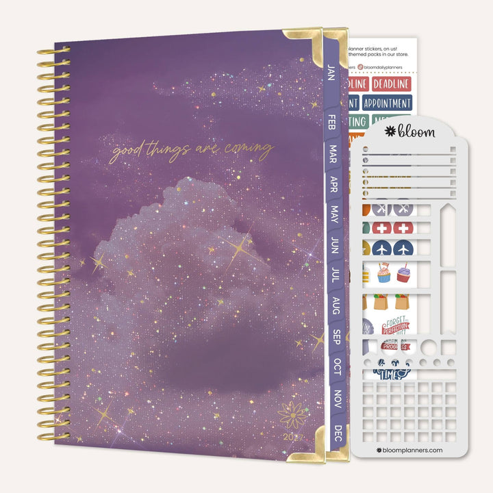 A spiral-bound planner with a purple galaxy cover, shown with a white sticker sheet and divider tabs on a light beige background. The cover reads 'good things are coming' in gold script, and the planner includes decorative stickers and gold spiral binding.