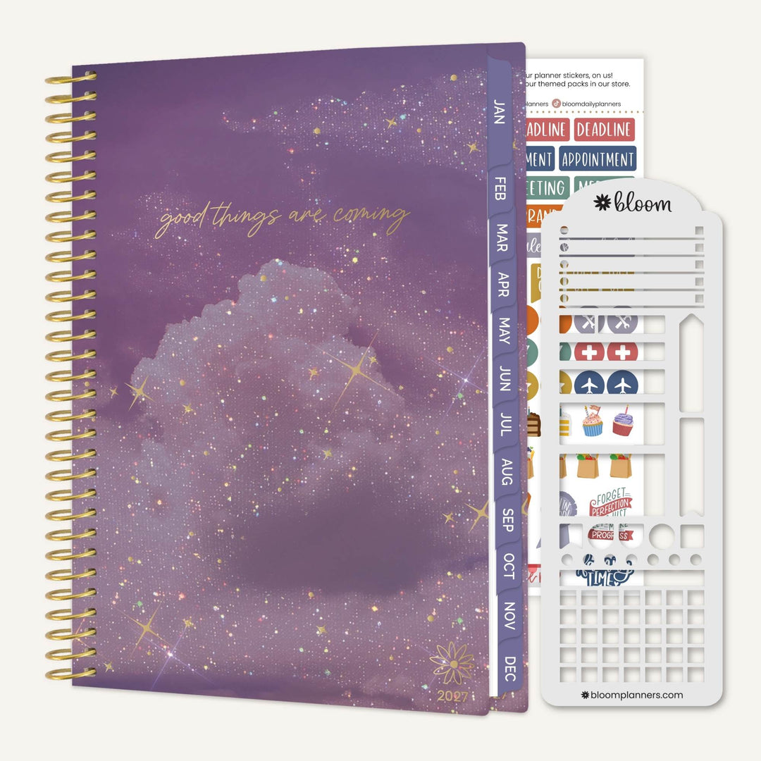 A spiral-bound planner shown with its decorative cover and included sticker sheet and divider pages on a light neutral background. The cover is purple with a cloudy night-sky print, gold script text reading 'good things are coming,' and gold spiral binding.