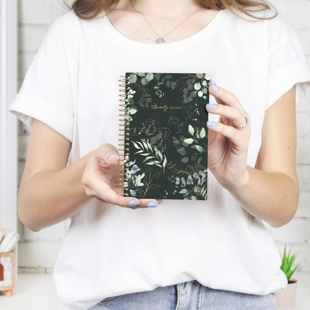 A person in a white t-shirt holding a spiral-bound notebook with a dark green botanical pattern and gold foil lettering that reads 'thou art mine'.