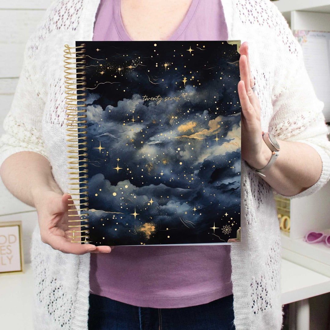 A person holding a spiral-bound notebook with a dark navy and black starry sky cover, featuring gold glitter clouds and stars, set against a bright indoor background.