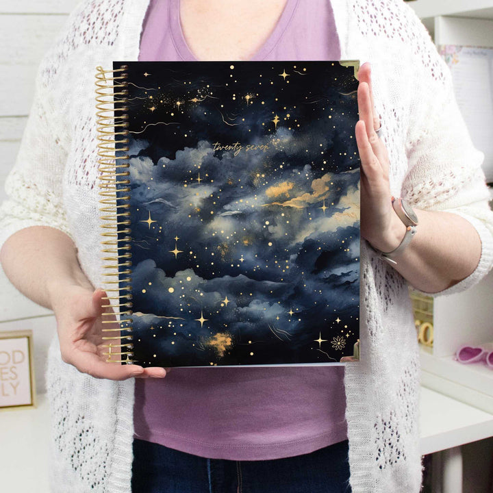 A person holding a spiral-bound notebook with a dark navy and black starry sky cover, featuring gold glitter clouds and stars, set against a bright indoor background.