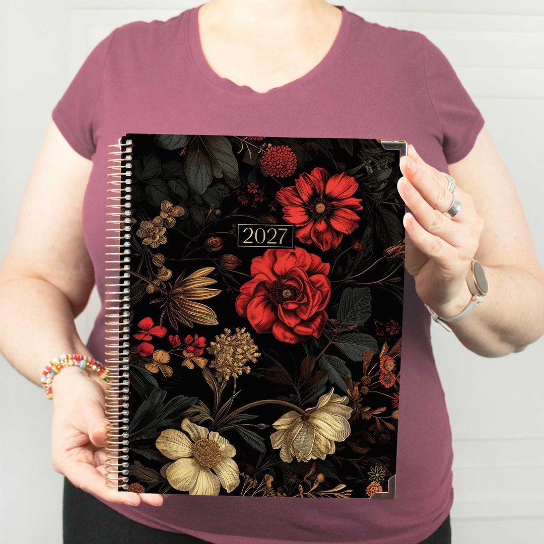 A person wearing a mauve t-shirt holds up a 2027 spiral-bound planner with a black cover decorated with red, beige, and gold floral illustrations.