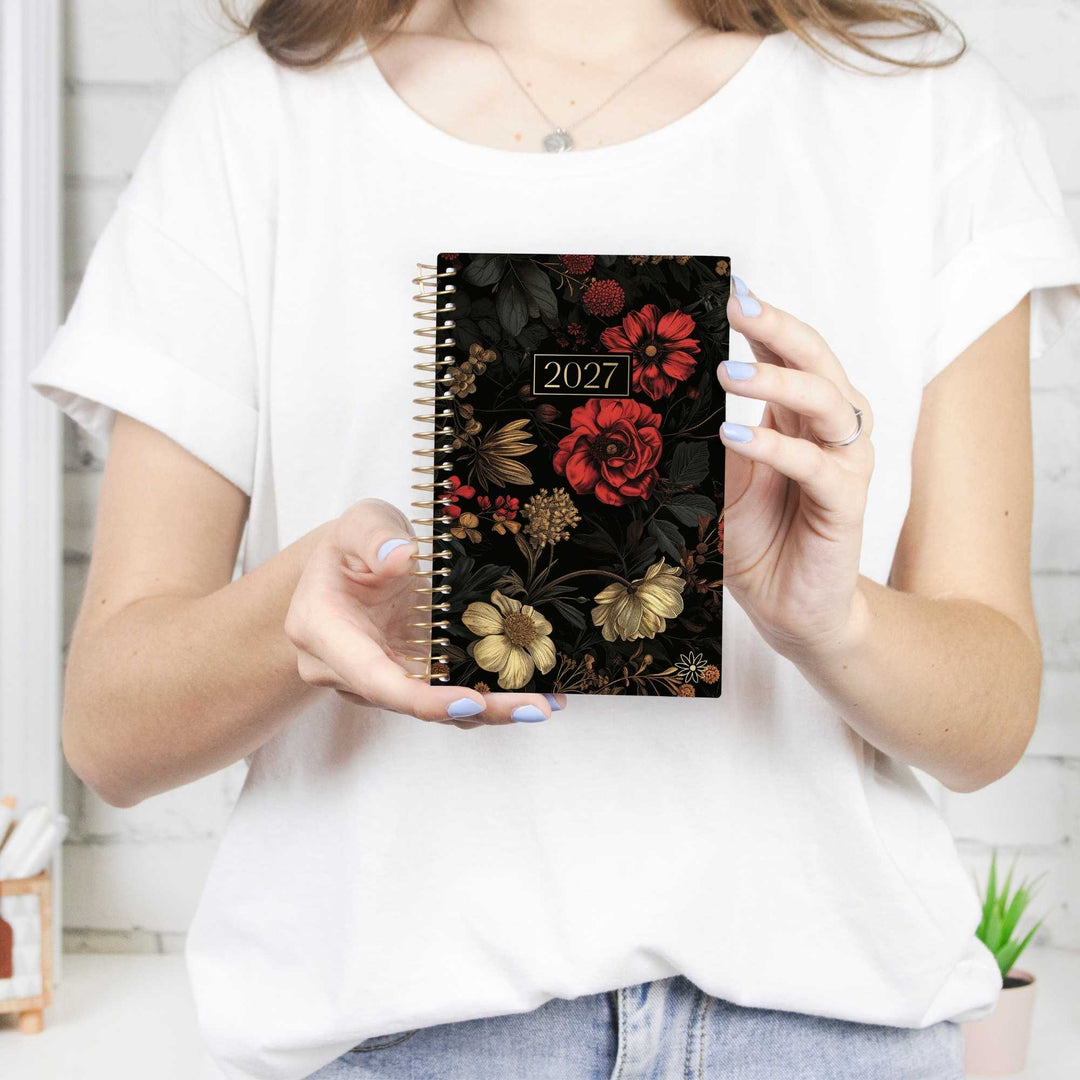 A woman in a white t-shirt holds a 2027 spiral-bound planner with a black background and a detailed red, gold, and brown floral pattern.