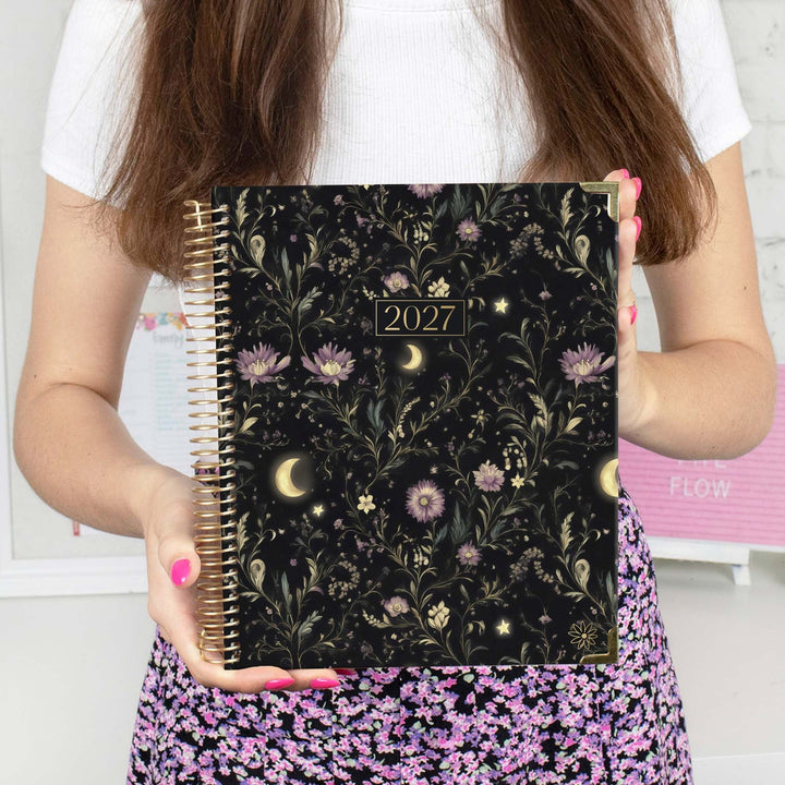 A person holds a 2027 spiral-bound planner with a black background and a gold-toned floral, moon, and star pattern. The person is wearing a white top and a purple and pink floral skirt, with pink fingernails.