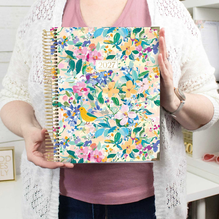 A person holding a 2027 spiral-bound planner with a colorful floral pattern featuring a yellow bird. The person is wearing a white knit cardigan over a dusty pink top and dark blue jeans, standing in a bright indoor setting.