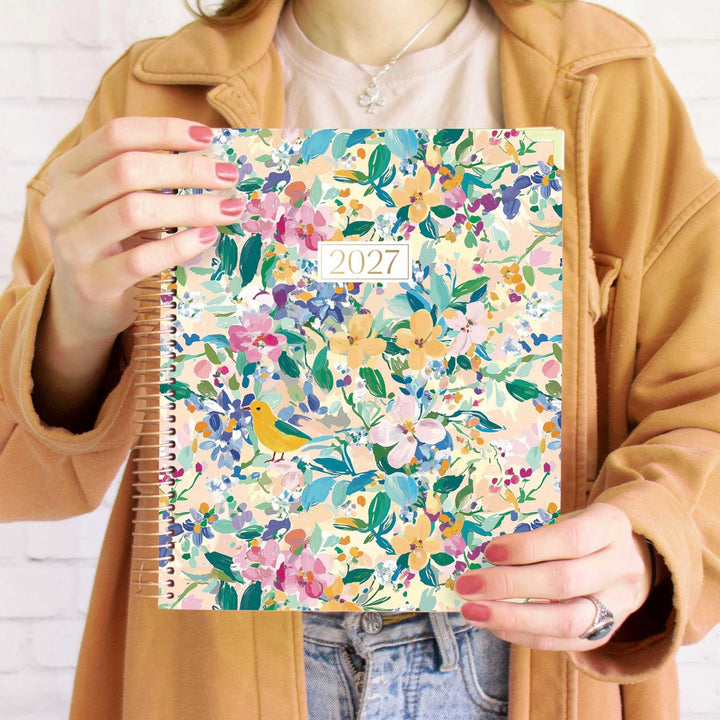 A person holding a 2027 spiral-bound planner with a colorful floral and bird pattern. The person is wearing a tan jacket over a light pink top and blue jeans, with pink nail polish and a silver necklace visible.