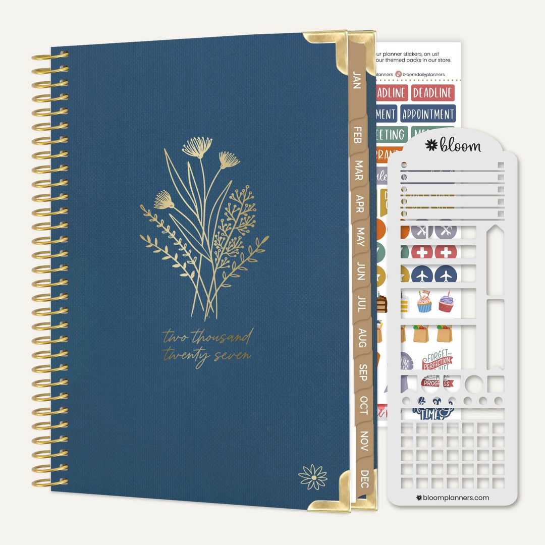 A navy blue spiral-bound planner with gold wire binding and gold floral line art on the cover, shown alongside a white sticker sheet and a gold divider tab. The sticker sheet has the brand name bloom visible.