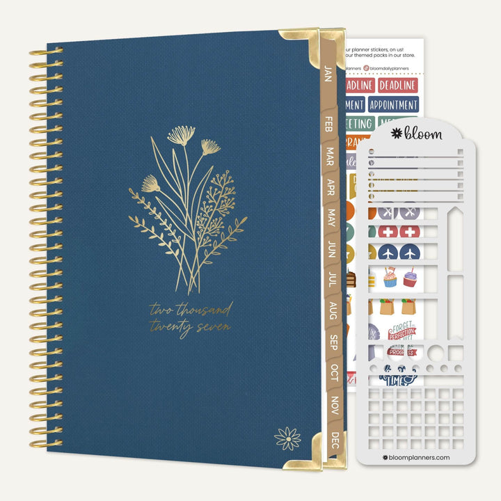 A navy blue spiral-bound planner with gold wire binding and gold floral line art on the cover, shown alongside a white sticker sheet and a gold divider tab. The sticker sheet has the brand name bloom visible.