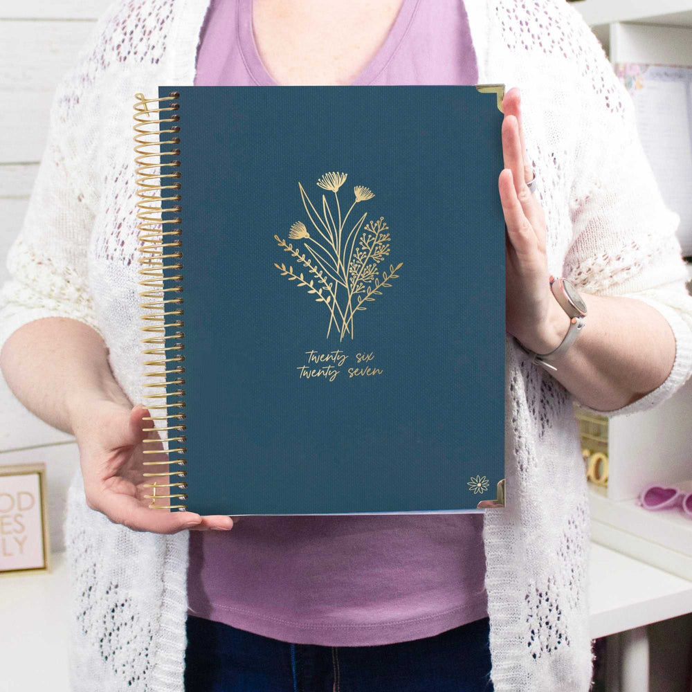 A person holding a navy blue spiral-bound planner with gold floral illustrations and the text 'twenty six twenty seven' on the cover, wearing a white knit cardigan over a lavender top. - hover
