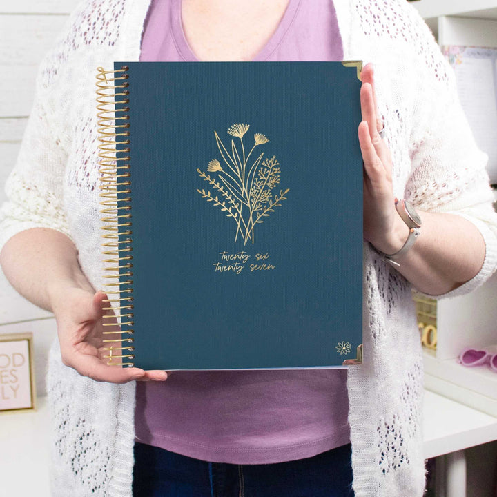 A person holding a navy blue spiral-bound planner with gold floral illustrations and the text 'twenty six twenty seven' on the cover, wearing a white knit cardigan over a lavender top.