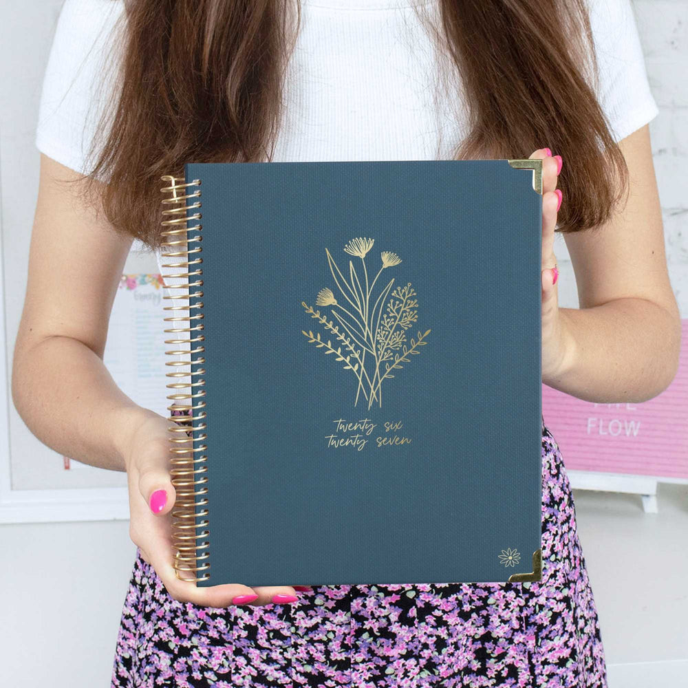 A person holds a navy blue spiral-bound planner or notebook with gold floral line art and the text 'twenty eight seven' on the cover. The person is wearing a white top and a black-and-purple floral skirt, with pink fingernails. - hover