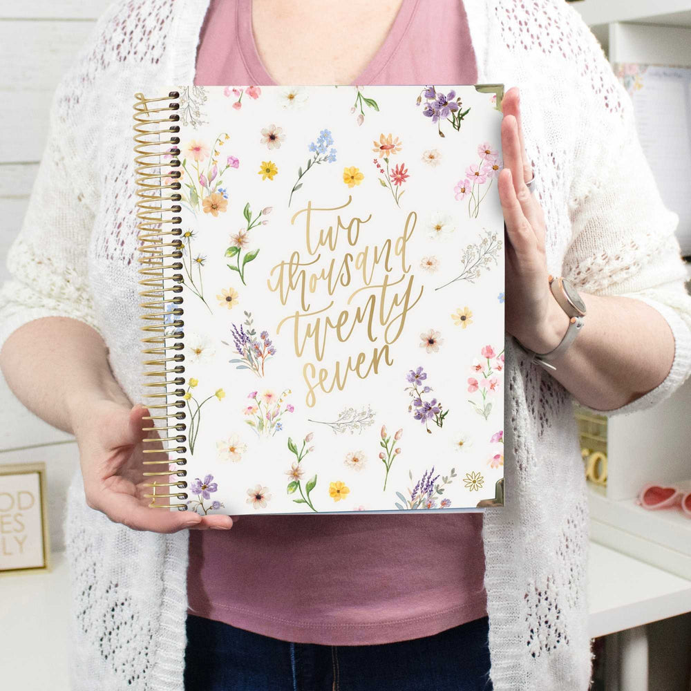 A person holding a white spiral-bound journal with gold foil text reading 'Two thousand twenty seven' and a floral border. The person is wearing a pink top and a white open-knit cardigan, standing in a bright indoor setting. - hover