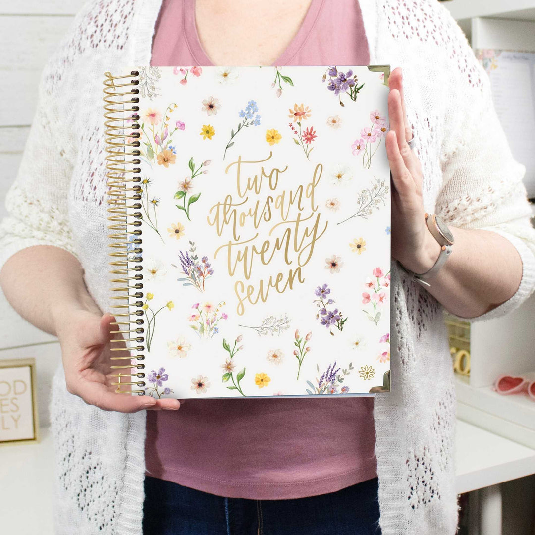 A person holding a white spiral-bound journal with gold foil text reading 'Two thousand twenty seven' and a floral border. The person is wearing a pink top and a white open-knit cardigan, standing in a bright indoor setting.