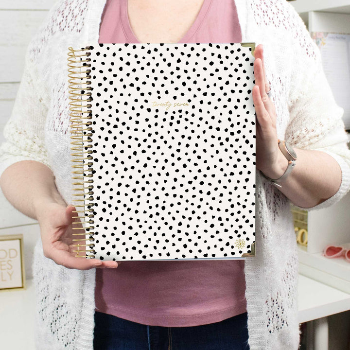 A person holding a 2027 spiral-bound planner with a beige and white checkerboard cover. The planner features a gold wire coil binding and a small gold sunflower logo in the bottom right corner.