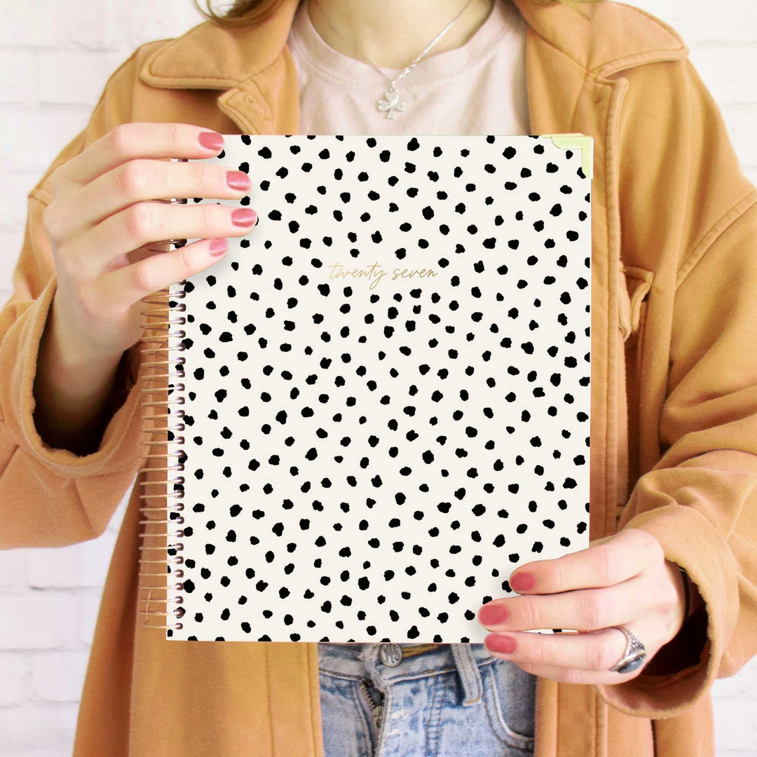 A person holding a white spiral-bound planner or notebook with a black polka dot pattern and gold foil text reading 'January 2025'. The person is wearing a tan jacket, a light pink top, and blue jeans, standing against a white brick wall.
