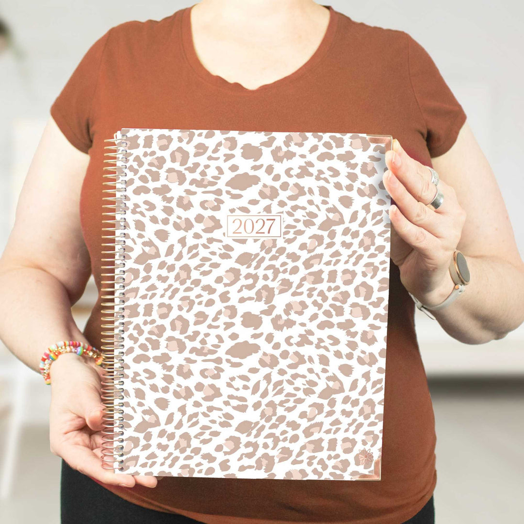 A person holding a 2027 spiral-bound planner with a leopard print cover. The person is wearing a brown t-shirt and holding the planner in front of their torso.
