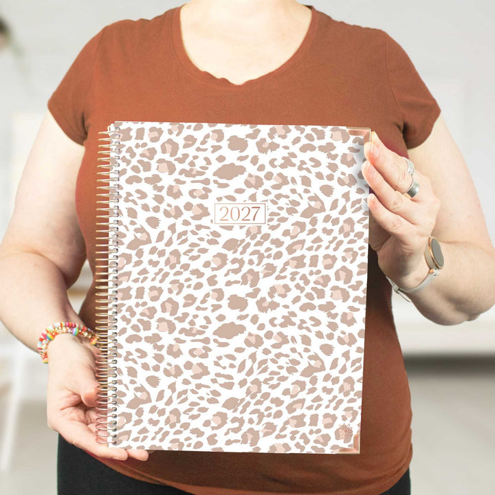 A person holding a 2027 spiral-bound planner with a leopard print cover. The person is wearing a brown t-shirt and holding the planner in front of their torso.