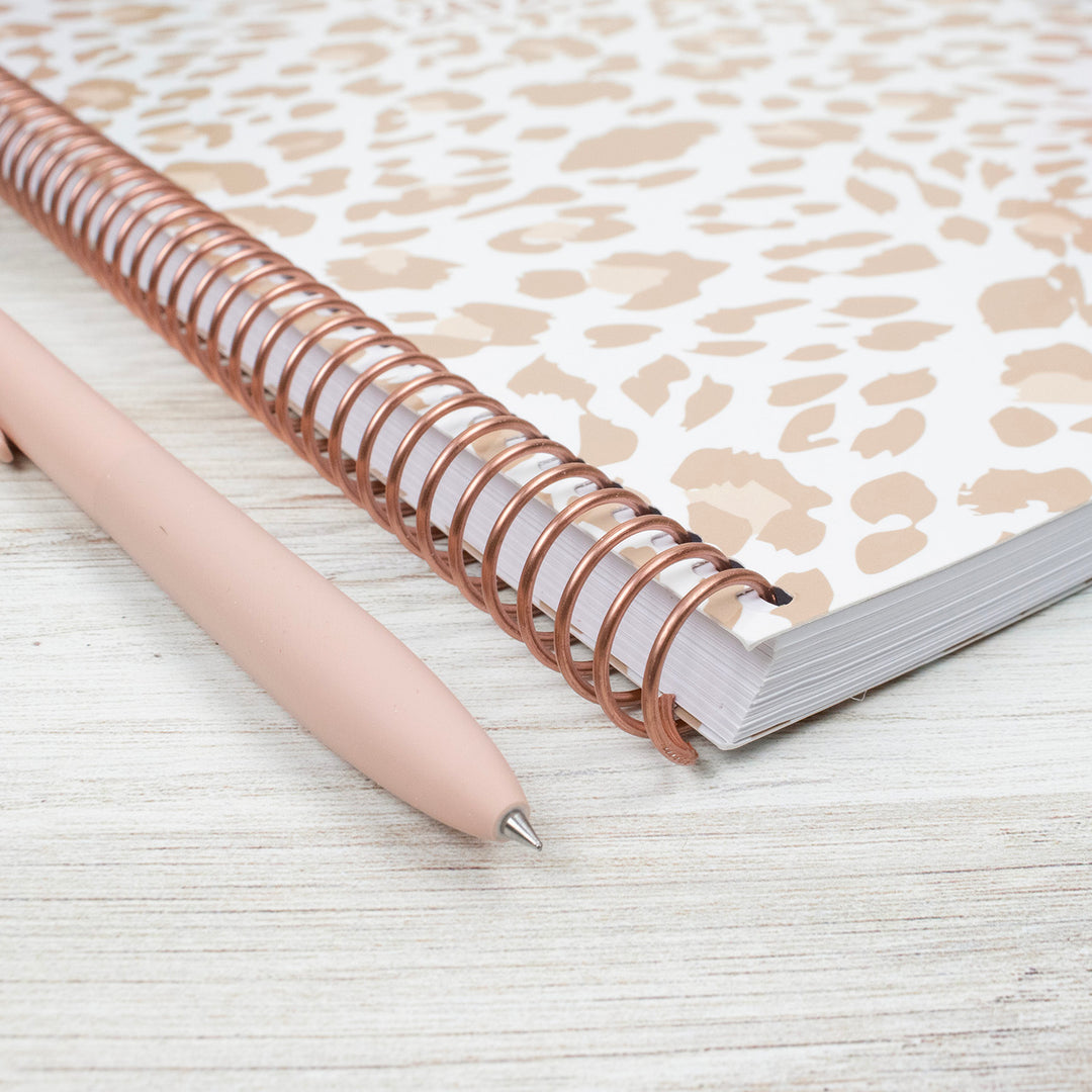 Planner with rose gold metal coil binding and a pen on a light wooden surface
