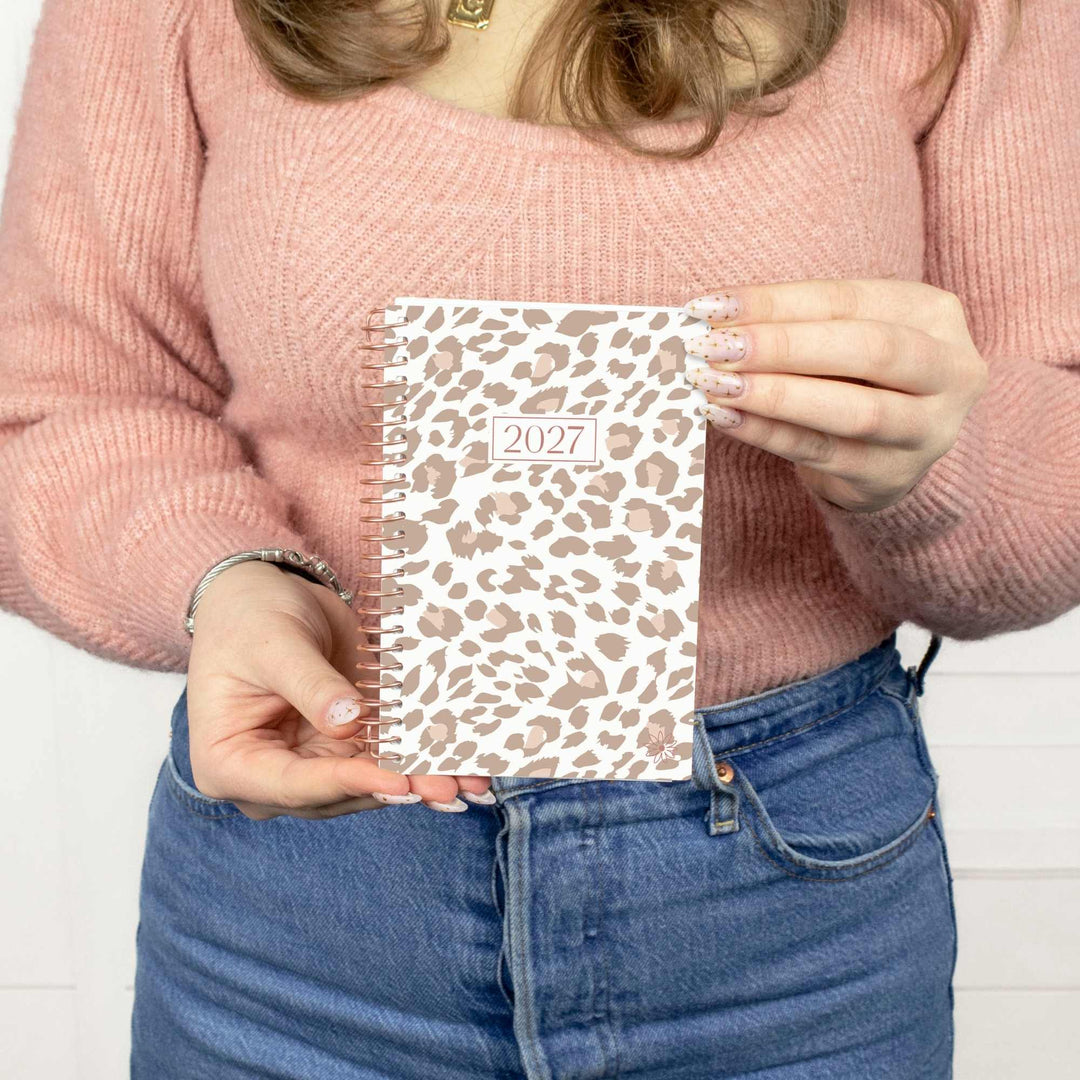 A person in a pink ribbed sweater and blue jeans holds a small spiral-bound 2027 planner with a beige and brown leopard print cover against a white paneled wall.