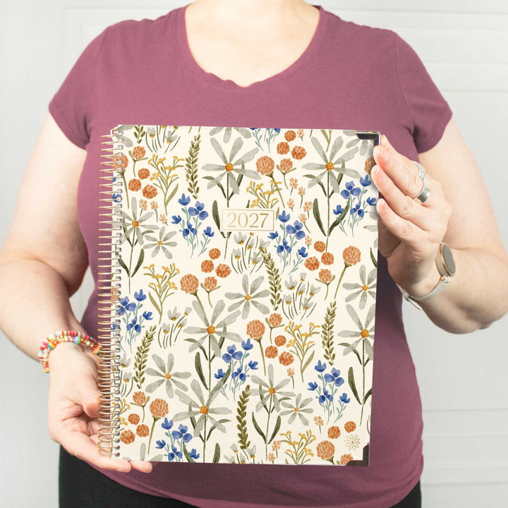 A person wearing a mauve t-shirt holds up a 2027 spiral-bound planner with a cream cover decorated with a colorful wildflower pattern.