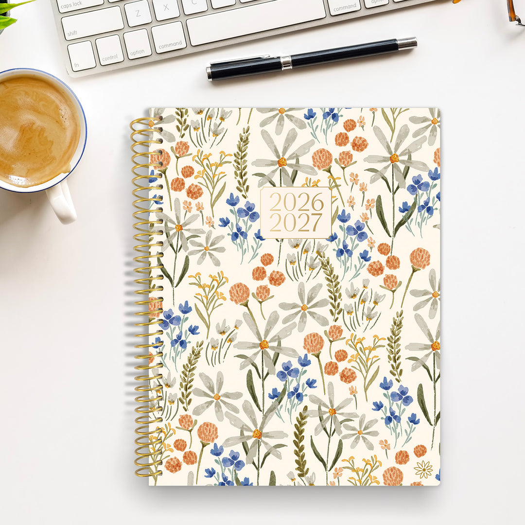 Floral-patterned planner for 2026-2027 on a desk with a cup of coffee and pen.