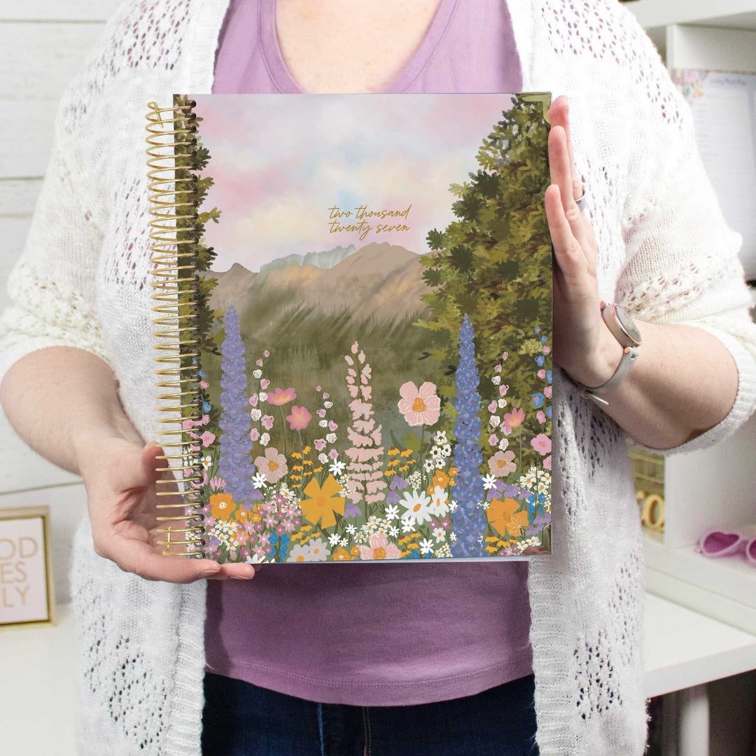 A person holding a spiral-bound planner with a colorful floral mountain landscape cover. The planner features the text 'two thousand twenty five' in gold script near the top.