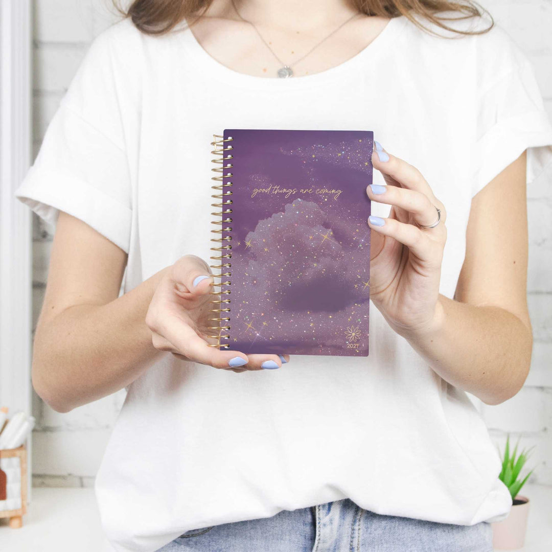 A person in a white t-shirt holds a purple spiral planner with a glittery cloud design and gold foil text that reads 'good thing everything is okay'.