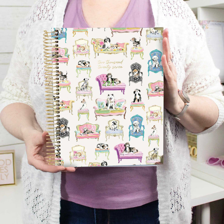 A person holds a spiral-bound planner or notebook with a white cover decorated with colorful illustrations of dogs lounging in pink, purple, and blue chairs. The person is wearing a white knit cardigan over a lavender shirt, with a watch on one wrist.
